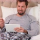 Smiling young man sitting on bed using his tablet in the bedroom 4K 4k