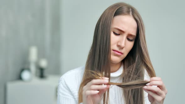 Upset Young Caucasian Woman Looking on Weak Unhealthy Dry Hair Ends at Home Interior alt