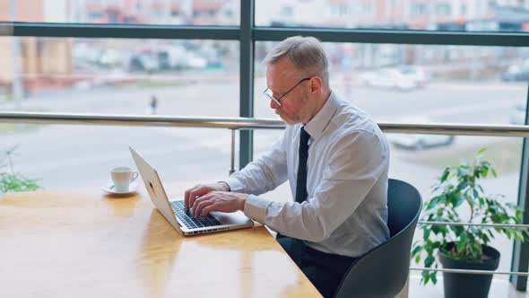 Mature man in white shirt sitting at the table near the window and working on a laptop alt