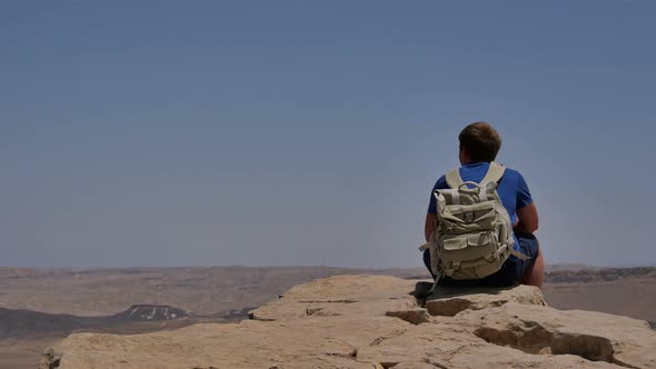 Young Man with Backpack Sitting on Cliff's Edge and Enjoying the Desert View