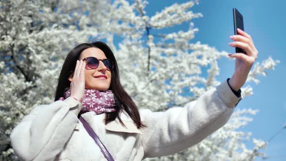Beautiful Smiling Woman Taking Selfie Using Smartphone Posing at White Flowering Sakura Tree alt