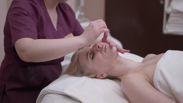 Beautiful Blond Caucasian Woman Enjoying Facial Massage Lying in Spa Salon with Unrecognizable alt