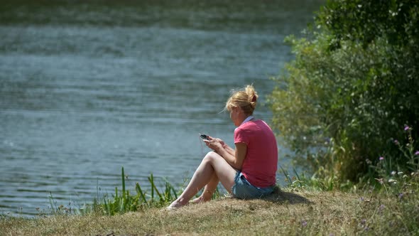 A young girl is sitting on the Bank of a river with headphones and listening to music.