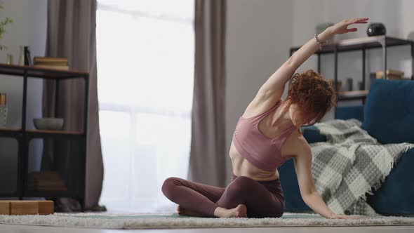 Adult Woman is Training Alone at Home Sitting on Floor in Living Room Stretching Hands and Torso alt