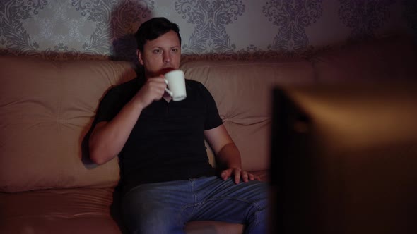 Young Man Watching Tv Sitting on the Couch with a Mug of Tea is Resting at Night alt