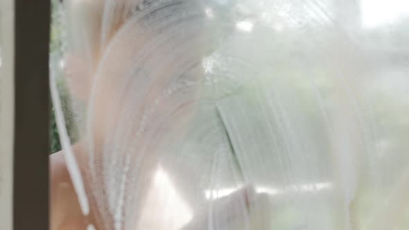 Boy Teenager with a Soaped Sponge Washes the Glass Near the Window From Dust alt