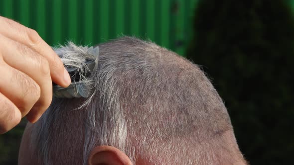 Cutting gray hair on the head of an elderly balding man with a cordless electric machine alt
