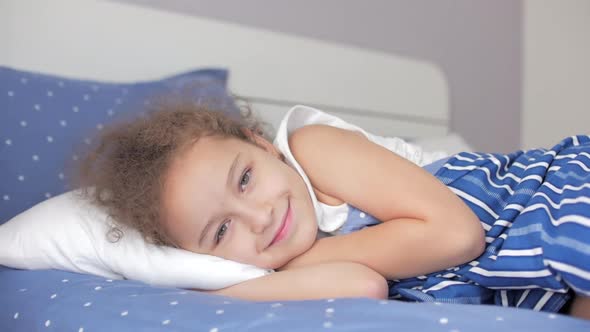 Portrait of a Happy Cute Funny Cheerful Little Child Girl in White Pajamas Wakes Up in Bed in the alt