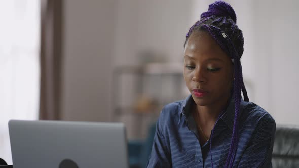 Portrait of Young Afroamerican Woman with Purple Dreadlocks at Table with Laptop Black Lady is Using alt