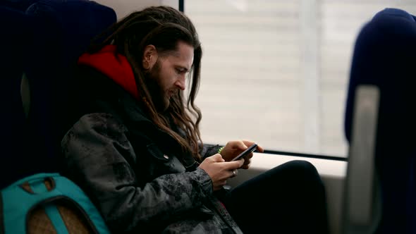Stylish Man with Long Hair is Riding Train and Surfing Internet By Smartphone with Wifi in Traincar alt
