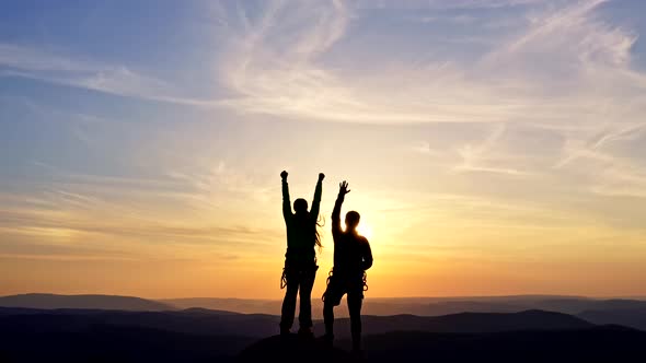 A Young Couple Cheerfully Raises Their Hands While Standing on a Mountaintop at Sunset