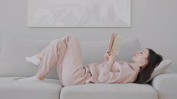 Young Caucasian woman with glasses reading book lying on her back on the sofa.