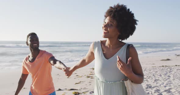 Smiling african american couple holding hands and walking on sunny beach alt