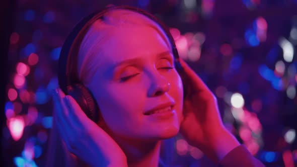Joyful Woman in Big Black Headphones Dances Against the Background of Flickering Lights
