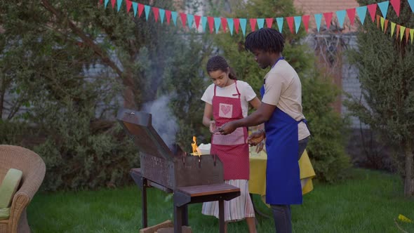Wide Shot of Concentrated African American Teenage Daughter Helping Father Setting Fire on Barbecue alt