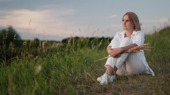 A Woman in White Clothes Rests on a Green Hill on a Warm Summer Evening alt