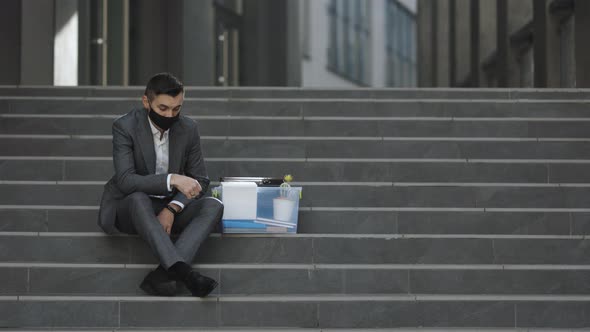 Fired Caucasian Male Office Worker in Medical Mask Sitting on Stairs in Depression With Box of Stuff