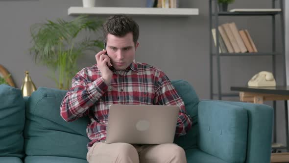 Angry Man with Laptop Talking on Smartphone on Sofa alt