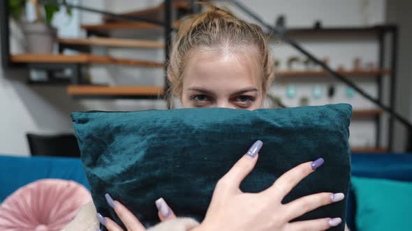 Beautiful Teenage Girl Covering Lower Face with Cushion Looking at Camera Sitting Indoors alt