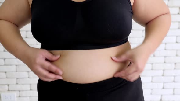 Overweight young woman in sportswear pinching her belly fat alt