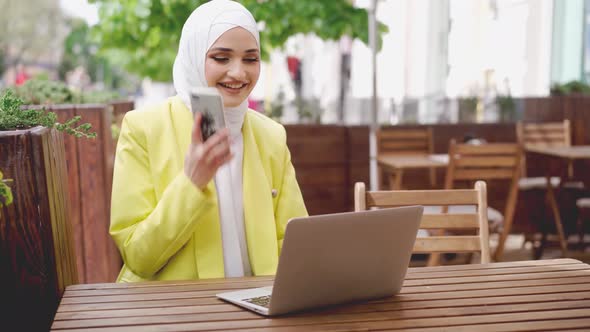 Smiling Young Muslim Woman in Headscarf Talking on the Phone and Using Laptop in Cafe alt