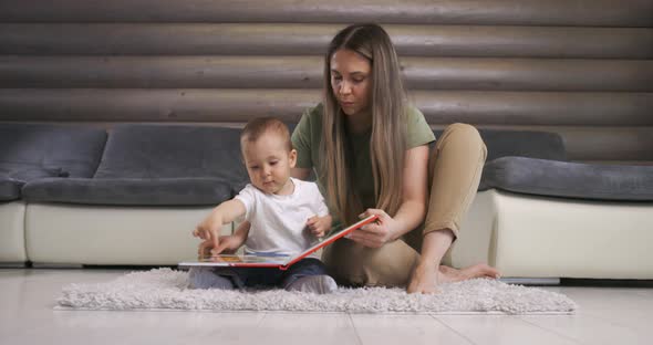Young Mother Reads a Book to Her Baby Son Who Listens with Curiosity While Sitting on Floor at Home