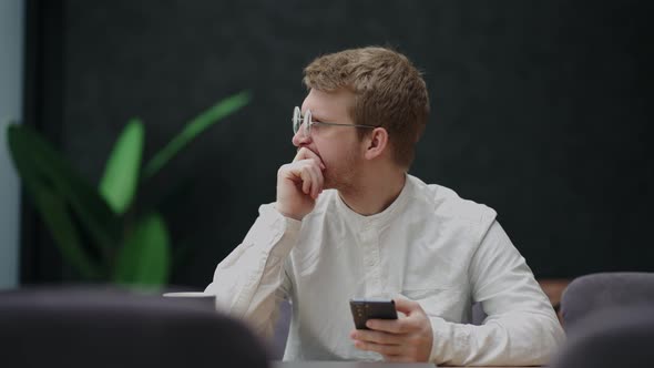 Handsome Pensive Man with Glasses and White Shirt is Sitting Alone in Cafe and Holding Smartphone in alt