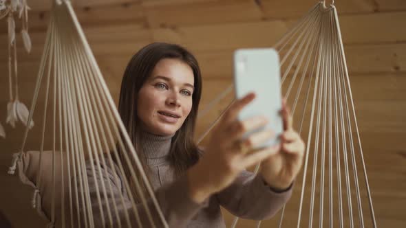 Portrait of Young Attractive Female in Stylish Outfit Making a Selfie on a Smartphone on a Swing in