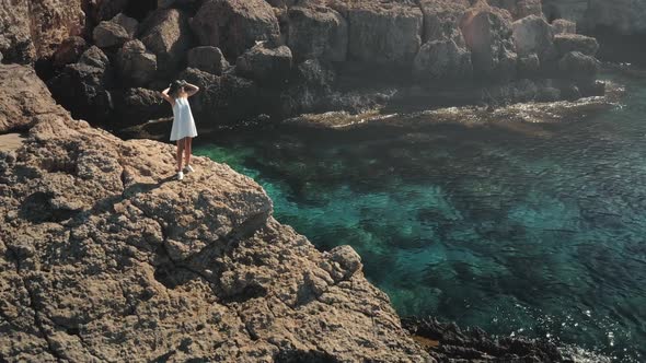 Hipster Woman in White Dress Standing on Rock Cliff Over Turquoise Sea Water Coast