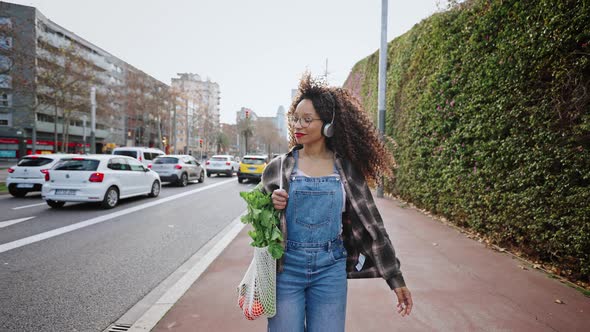 Lovely Student Walks Along a City Street at Dawn with a Bag of Oranges and Herbs and Listens to alt