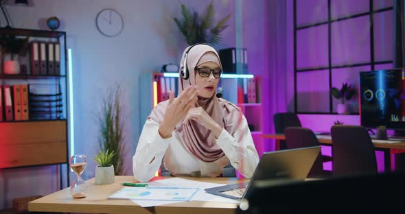 Arabian Businesswoman in Hijab Wears Headset During Online Conference on Laptop in the evening alt