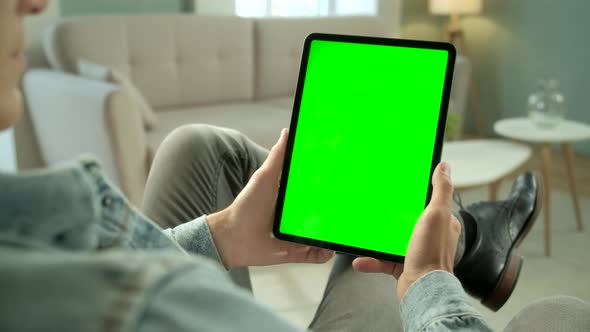 Over The Shoulder View Young Man at Home Sitting on a Chair Using With Green Mock-up Screen Tablet alt
