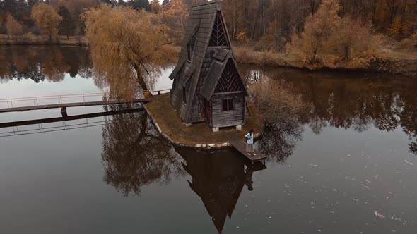 Aerial View of Girl Traveler Standing By Old Wooden House Chalet By Lake Among Trees at Forest with alt