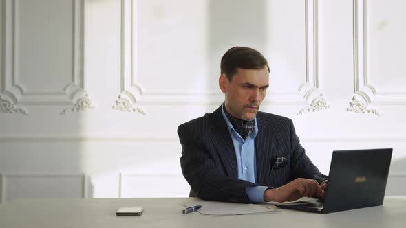 Man Works at a Laptop in a Suit Sitting at a Table in a Large Hall