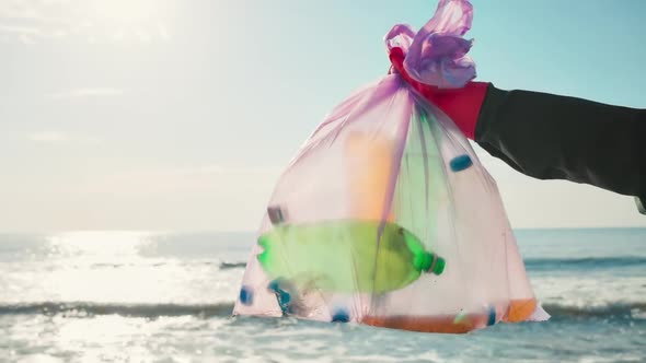 A volunteer in protective gloves holds a plastic bottle and puts it in a garbage bag alt