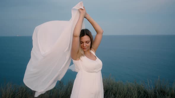 Pretty Woman Stands on Cliff Over Sea and Holds Long White Fabric Veil, Like Sails Fluttering in alt