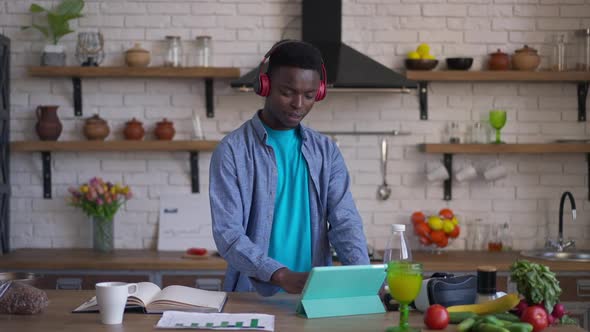 Handsome Young Man in Headphones Scrolling Tablet Screen Showing Thumb Up Looking at Camera with alt