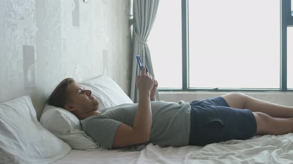 Young Handsome Man Lies in Bed By the Window Uses the Phone