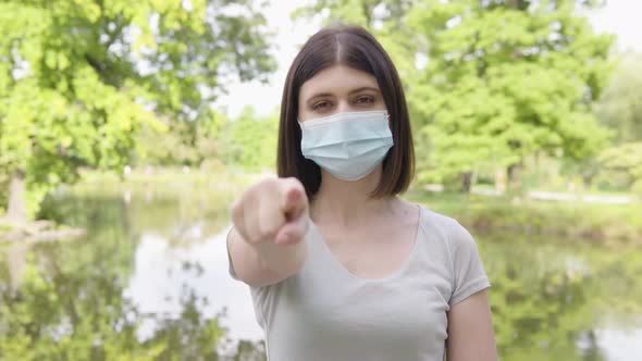 A Young Caucasian Woman in a Face Mask Points at the Camera and Nods in a Park alt