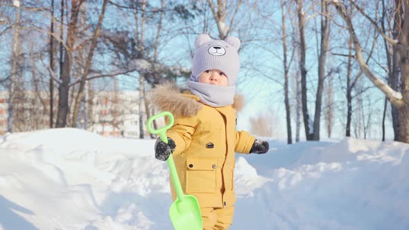 A baby of 15-23 months on a walk in a winter park walks along a snow-covered path. Toddler waves to alt