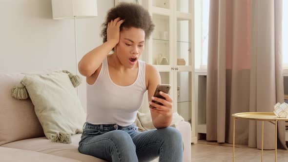 Surprised young African American woman shocked using the phone to receive an online win notification alt