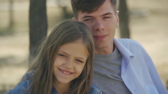 Portrait of Cute Little Girl Happily Embracing with Love and Care Her Elder Teenage Brother in Park alt