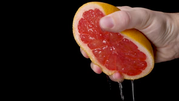 Squeezing out a red juicy grapefruit with a white hand in front of a black background. alt