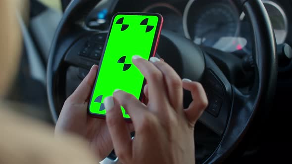 Close-up of a Woman's Hand with a Green Phone Screen on the Steering Wheel of a Car. A Young Driver alt