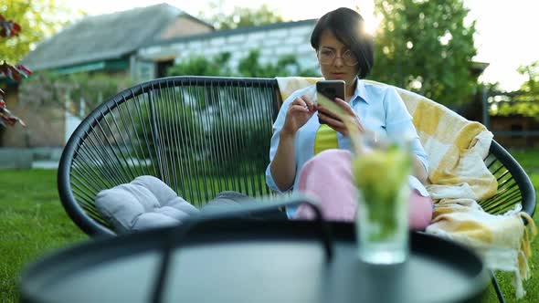 Woman in Glasses Using Smart Phone Sitting on Chair at Backyard Outdoor at Home with Lemonade
