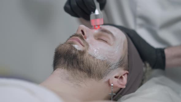 Closeup Face of Male Client Lying on Beauty Bed As Beautician in Gloves Doing Red LED Light alt