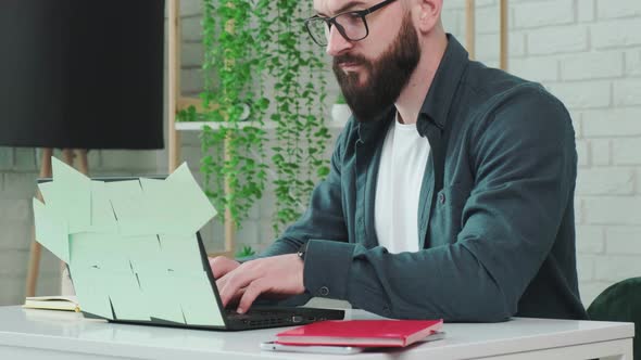 Bearded Man Types at the Laptop and Write Notice on the Paper Notebook alt
