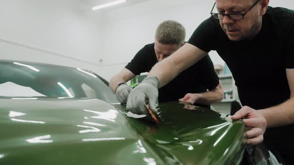 Two Men are Vinyl Wrapping a Car in Dark Green Color Using Plastic Cards alt