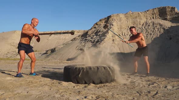 Two Male Athletes Training Together Hit the Wheel with a Hammer at Sunset in the Mountains on the alt