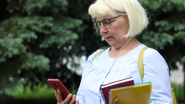 Adult Businesswoman in Eyeglasses Uses a Smartphone While Standing on the Street in a City Park